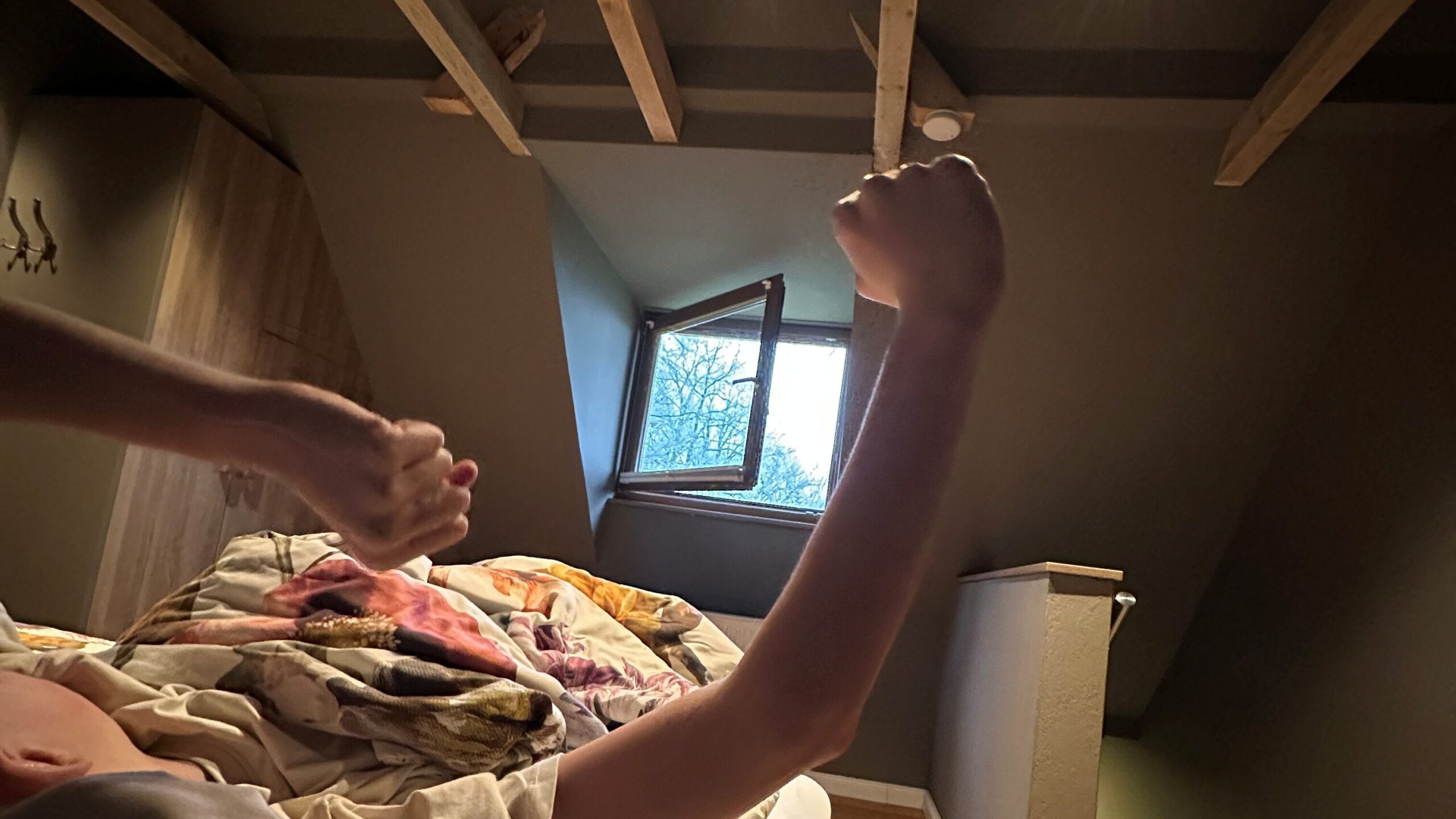 Entspannter Start in den Tag - Kind streckt sich morgens im Bett unter der Dachschräge mit offenem Fenster in der Ferienwohnung.