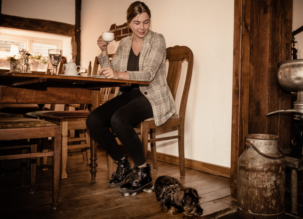 Frau mit Kaffeetasse sitzt im Hofcafé von Bussjans Hof, Hund liegt zu ihren Füßen.