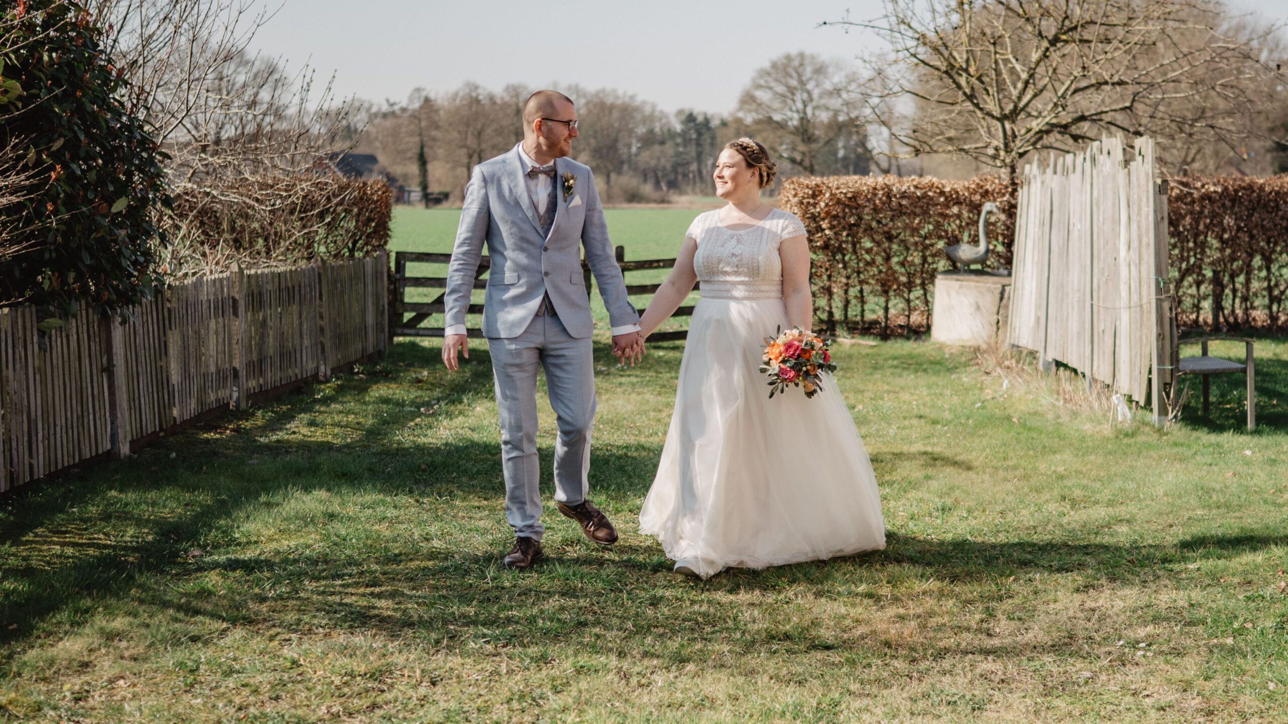 Ein Brautpaar spaziert Hand in Hand über eine grüne Wiese vor der ländlichen Kulisse mit Holzzaun und Bäumen - idealer Ort für romantische Fotoshootings im Fachwerkstil.